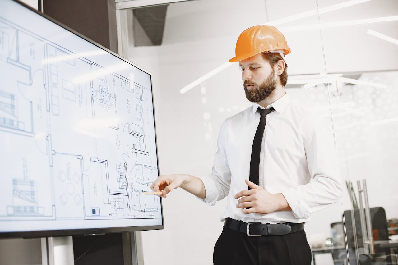 Architect in hard hat reviewing blueprints on a screen, discussing plans in modern office.